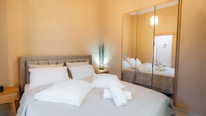 Una cama bien hecha con ropa de cama blanca, una mesita de noche, un espejo y una lámpara de pared.