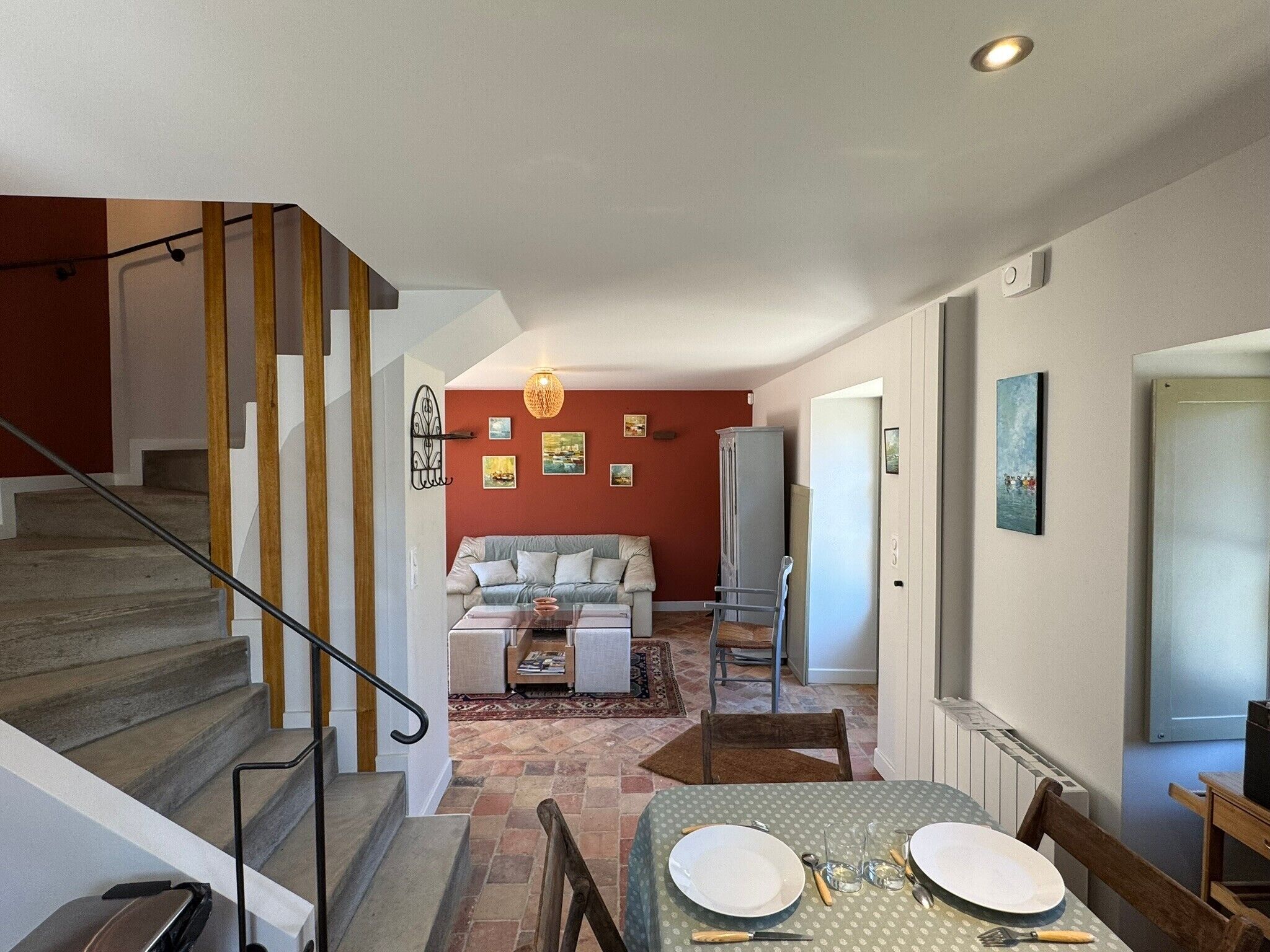 Un escalier mène à un espace de vie comprenant un canapé, une table à manger dressée avec de l’assiette et des couverts, ainsi qu’un couloir avec une porte et un tableau.