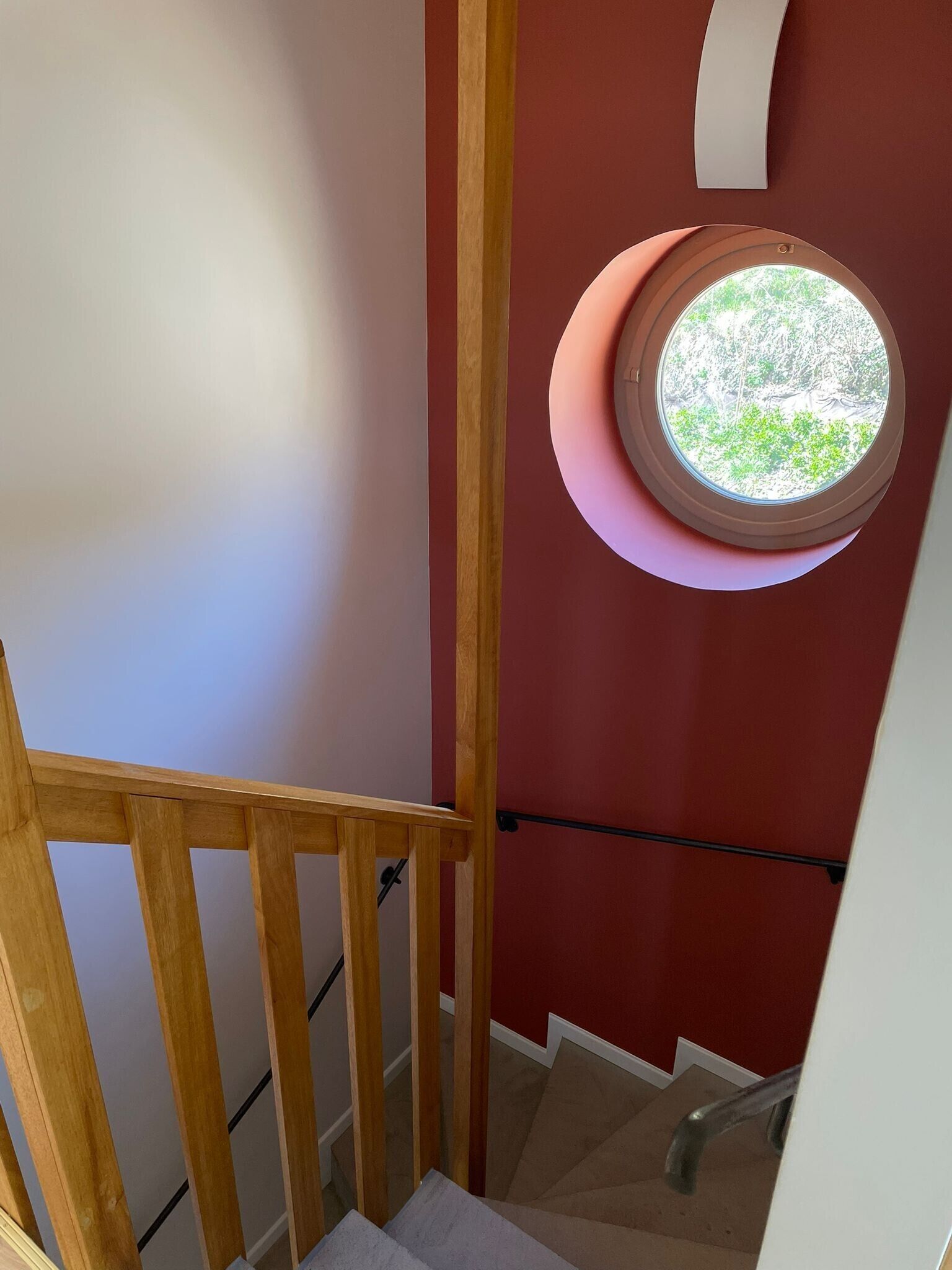 Una scala con ringhiera in legno, un muro rosso e una finestra circolare che si affaccia sul verde.
