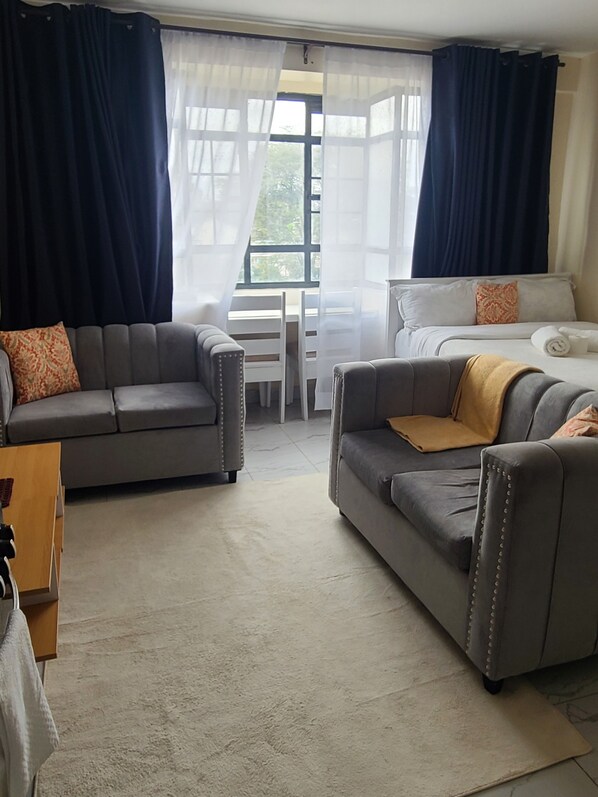 Ein Zimmer mit zwei grauen Sofas, einem Bett mit weißer Bettdecke, einem Holznachttisch und Blick durch ein Fenster mit durchscheinenden Vorhängen.