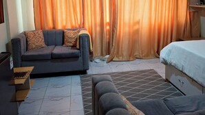 Et hotelværelse med en sofa, en seng, et skrivebord med en bærbar computer og et gardin.