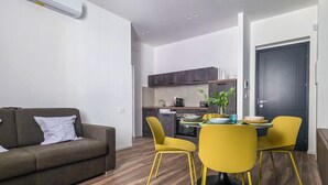 A modern living room with a sofa, dining table, and chairs, a kitchen in the background, and a door. - Grey Pearl Suites - Green Pearl (Manfredonia)