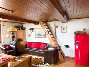 A cozy living room with a wooden ceiling, a red refrigerator, a brown leather sofa with red cushions, a wooden table with a cowhide rug, and a staircase.