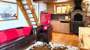 En mysig stuga med träpanel i taket, en soffa med röda kuddar, en läderfåtölj med en kudde prydd med Schweiz flagga, ett köksutrymme med träskåp, ett rött kylskåp och en matta gjord av kohud.