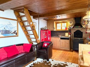A cozy cabin interior with a wooden ceiling, a sofa with red cushions, a leather chair with a Swiss flag pillow, a kitchen area with wooden cabinets, a red refrigerator, and a cowhide rug.