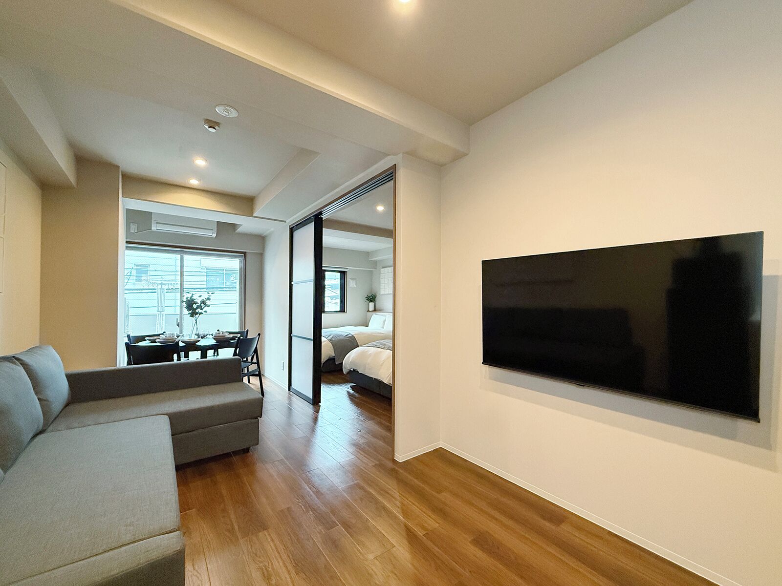 Una sala moderna con un televisor de pantalla plana grande, un sofá, un comedor con mesa y sillas, y un dormitorio con cama y puerta corredera.