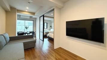 Una sala moderna con un televisor de pantalla plana grande, un sofá, un comedor con mesa y sillas, y un dormitorio con cama y puerta corredera.