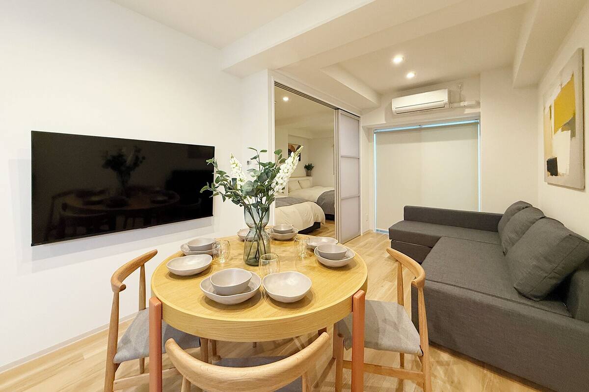 Una sala moderna con una mesa de comedor para cuatro personas, un sofá, un televisor montado en la pared y una puerta corrediza que da a otra habitación.