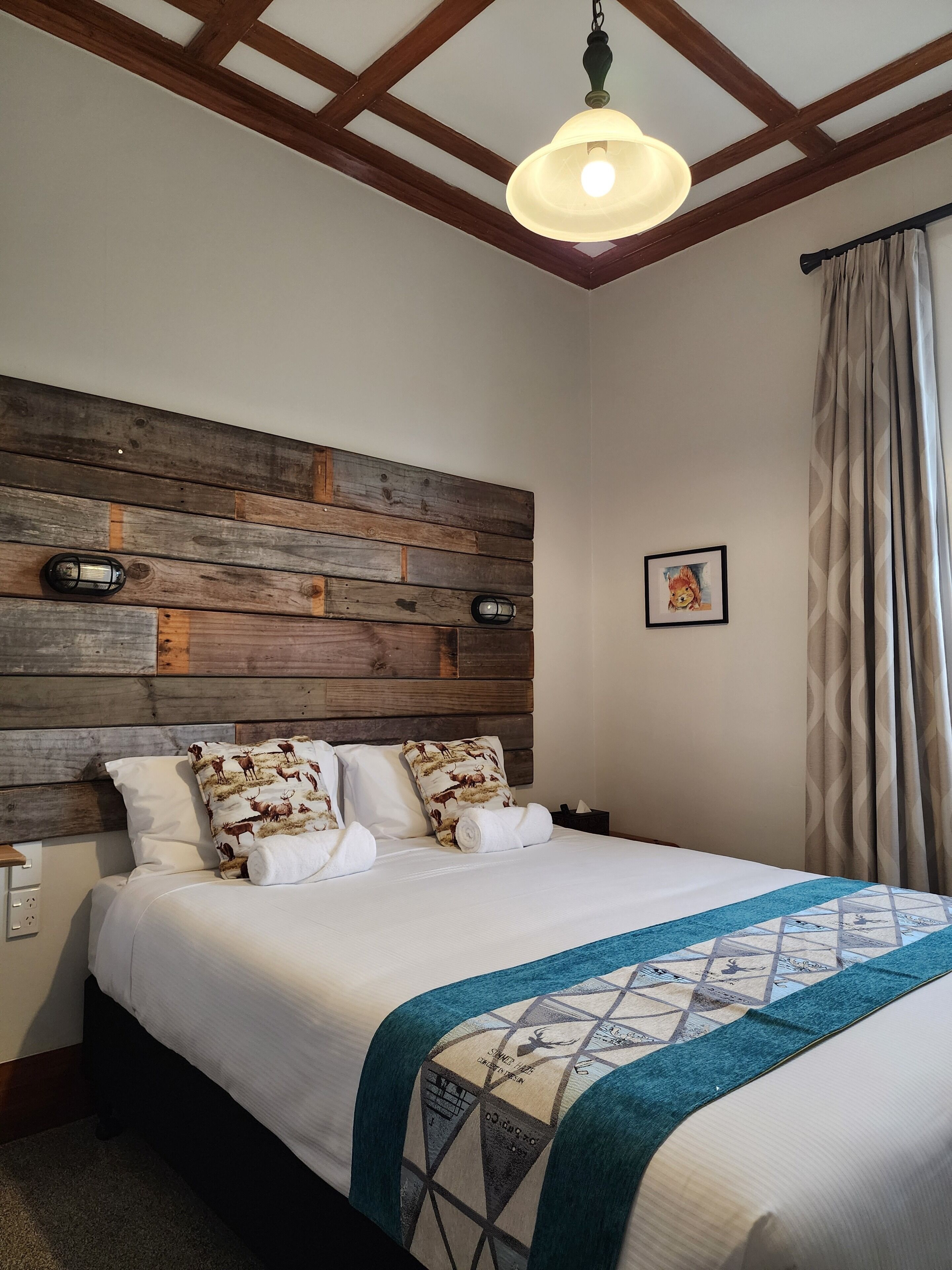 Une chambre à coucher avec une tête de lit en bois, un lit recouvert de draps blancs, une couverture turquoise et un luminaire suspendu.