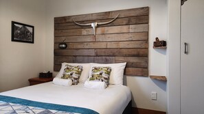 A bed with a wooden headboard, two pillows with a nature print, a bedside table with a lamp, and a framed picture on the wall.
