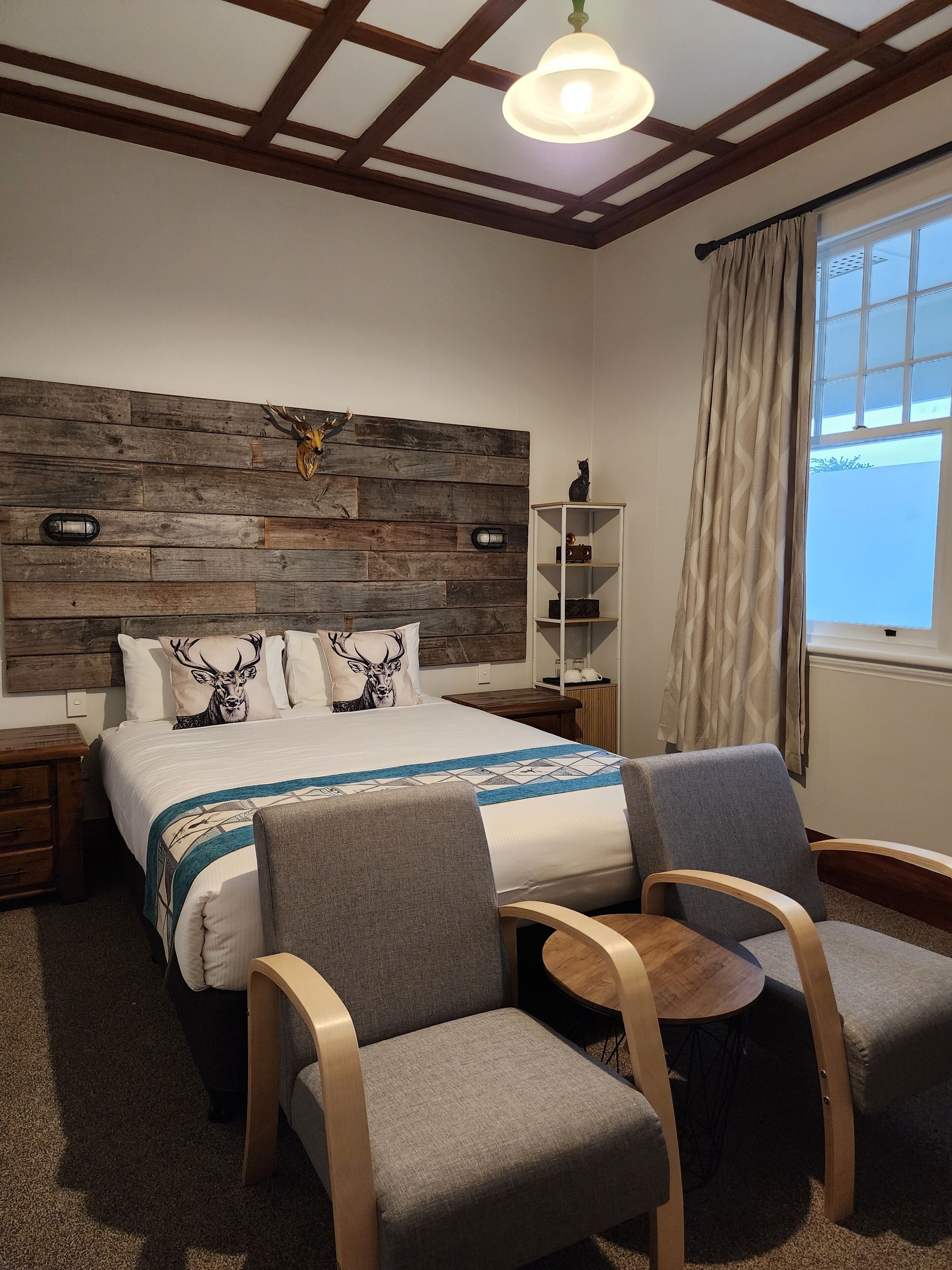 Un dormitorio con cabecera de madera, cama con almohadas estampadas, mesita de noche, una silla, una pequeña mesa y una ventana con cortinas.