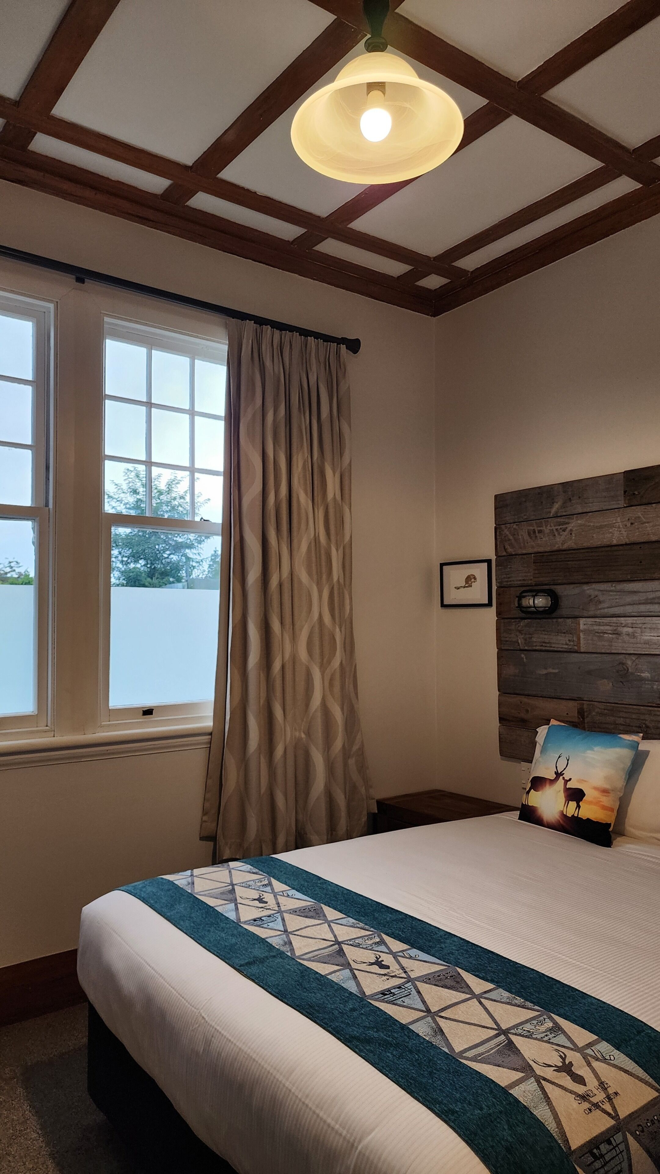 A bedroom with a bed, a window with curtains, a wooden headboard, and a wall-mounted light fixture.