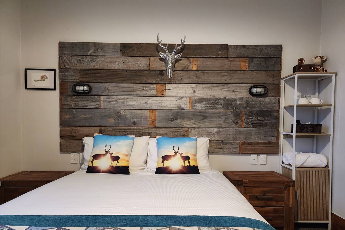 Une chambre avec une tête de lit en bois, deux oreillers ornés de motifs de cerfs, un lit recouvert d’une couverture à motifs géométriques, une table de chevet, une étagère avec des articles de toilette et un tableau accroché au mur.
