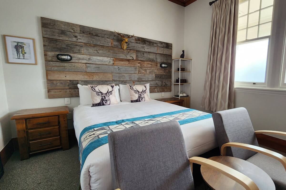 Une chambre avec une tête de lit en bois, un lit avec une literie à motifs, une table de chevet, une chaise et une fenêtre avec des rideaux.