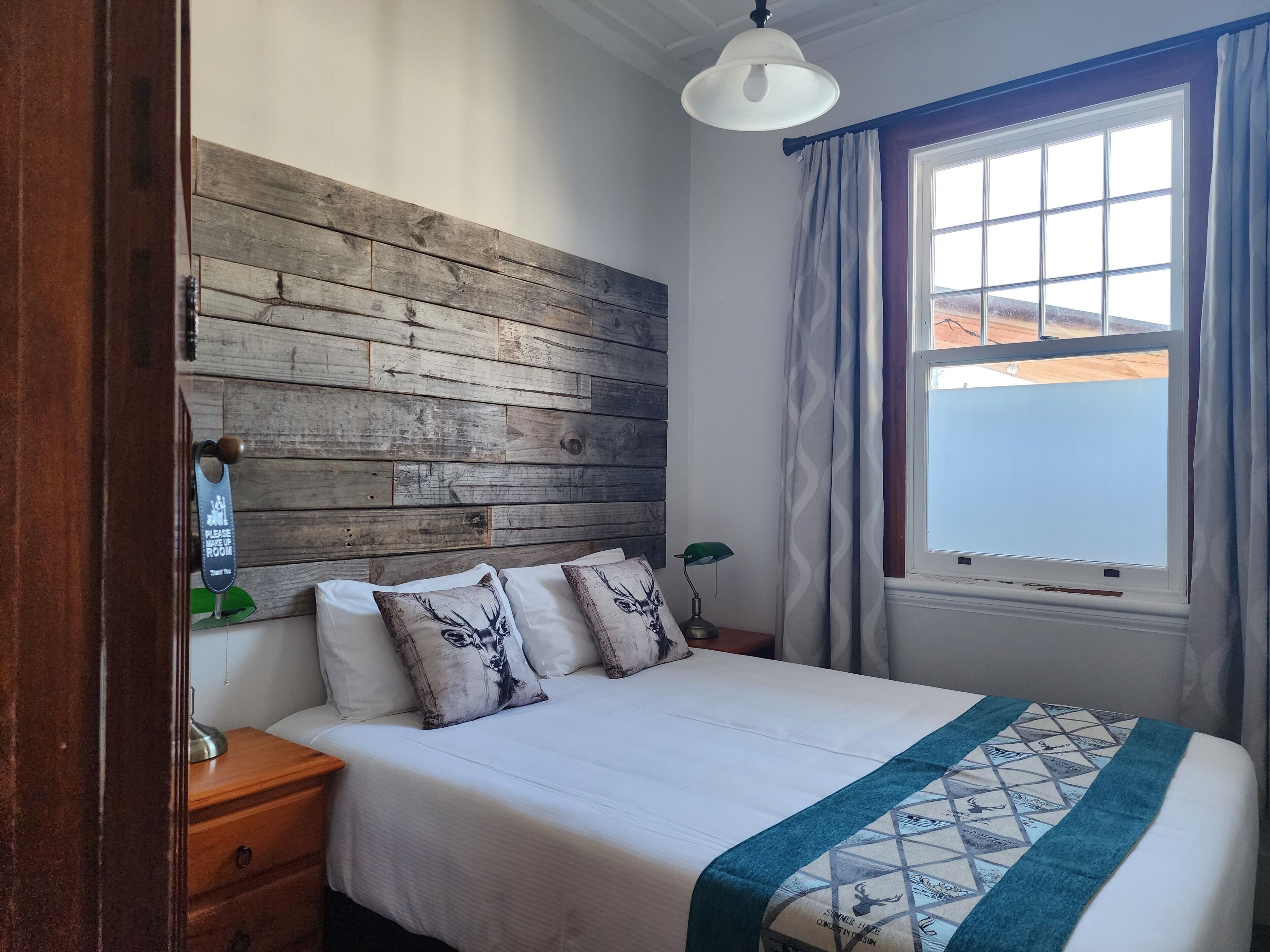 Un dormitorio con cabecera de madera, cama con ropa de cama blanca y almohadas estampadas, lámpara de noche y ventana con cortinas.