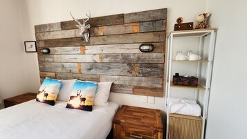 Un dormitorio con cabecera de madera, dos almohadas estampadas con ciervos, una mesita de noche y un estante blanco con objetos.