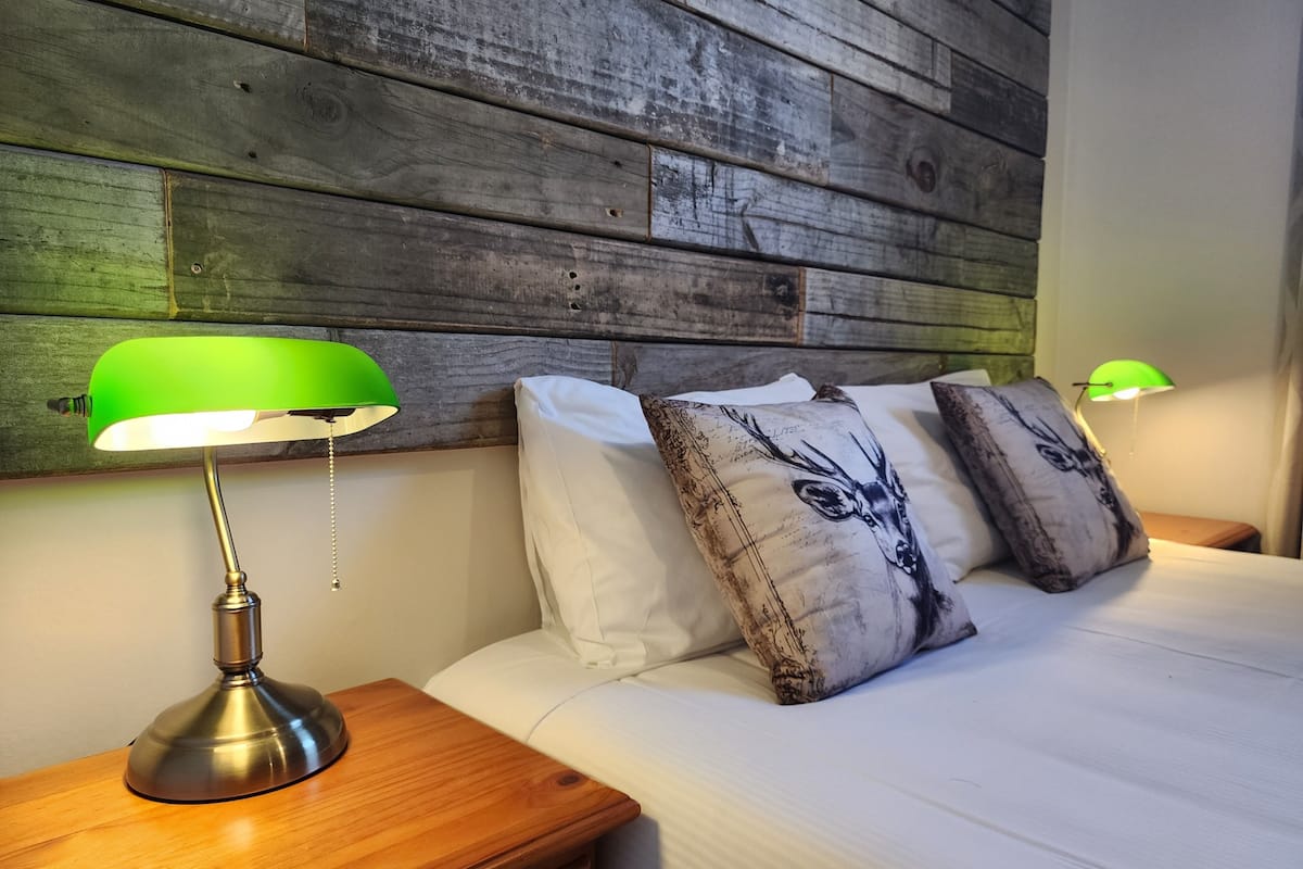 Un lit bien fait, avec du linge de lit blanc, deux lampes vertes posées sur des tables de chevet en bois, et une tête de lit en bois.