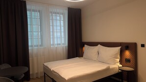 A hotel room with a bed, a round table, a chair, and a window with curtains. - Factory Suites (Prague)