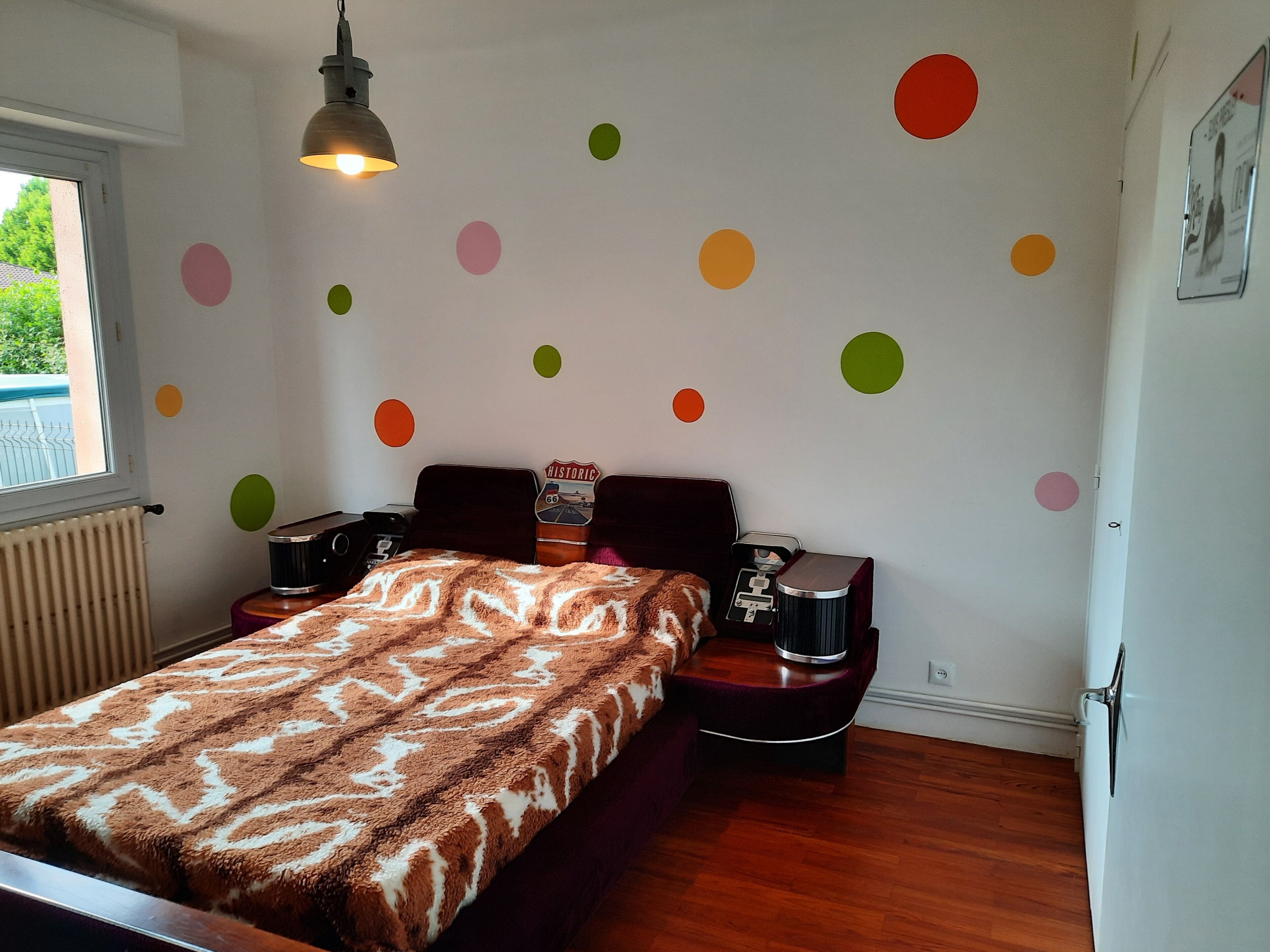 Un dormitorio con cama, mesitas de noche, cómoda, radiador, ventana y una pared decorada con pegatinas circulares de colores.