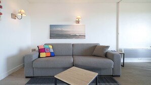Un soggiorno moderno con un divano grigio, un cuscino decorativo colorato, un tavolino da caffè in legno e un quadro raffigurante una spiaggia.