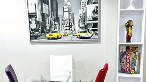 A modern room with a glass table, two chairs, a bookshelf, and a cityscape painting. - Private, Cozy, Luxury, Spacious One Bedroom Suite (Airdrie)