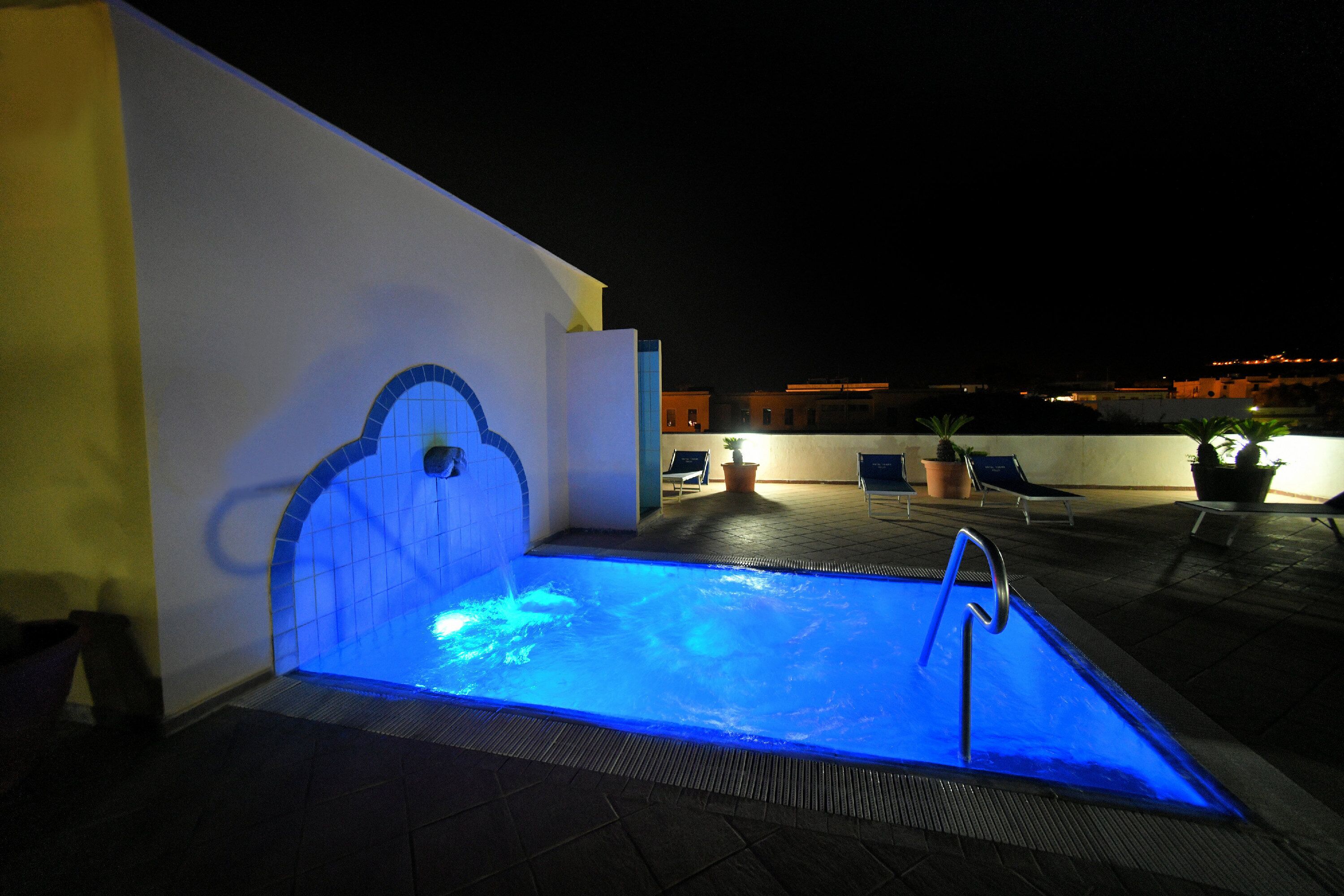 Una piscina exterior con un borde iluminado en azul, una escalera metálica y sillones de descanso al fondo.