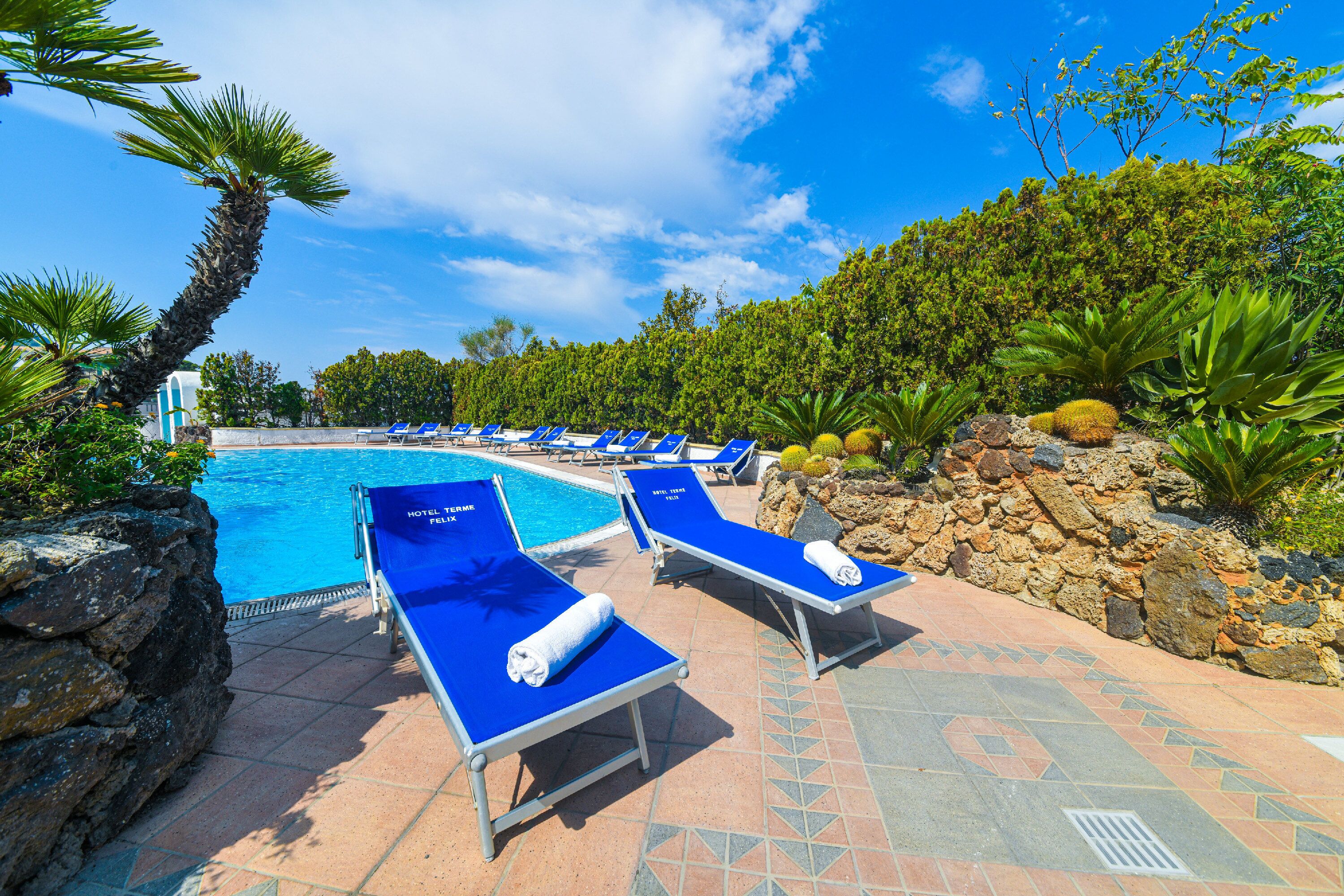 Uma área à beira da piscina com espreguiçadeiras, piscina e vegetação ao redor.