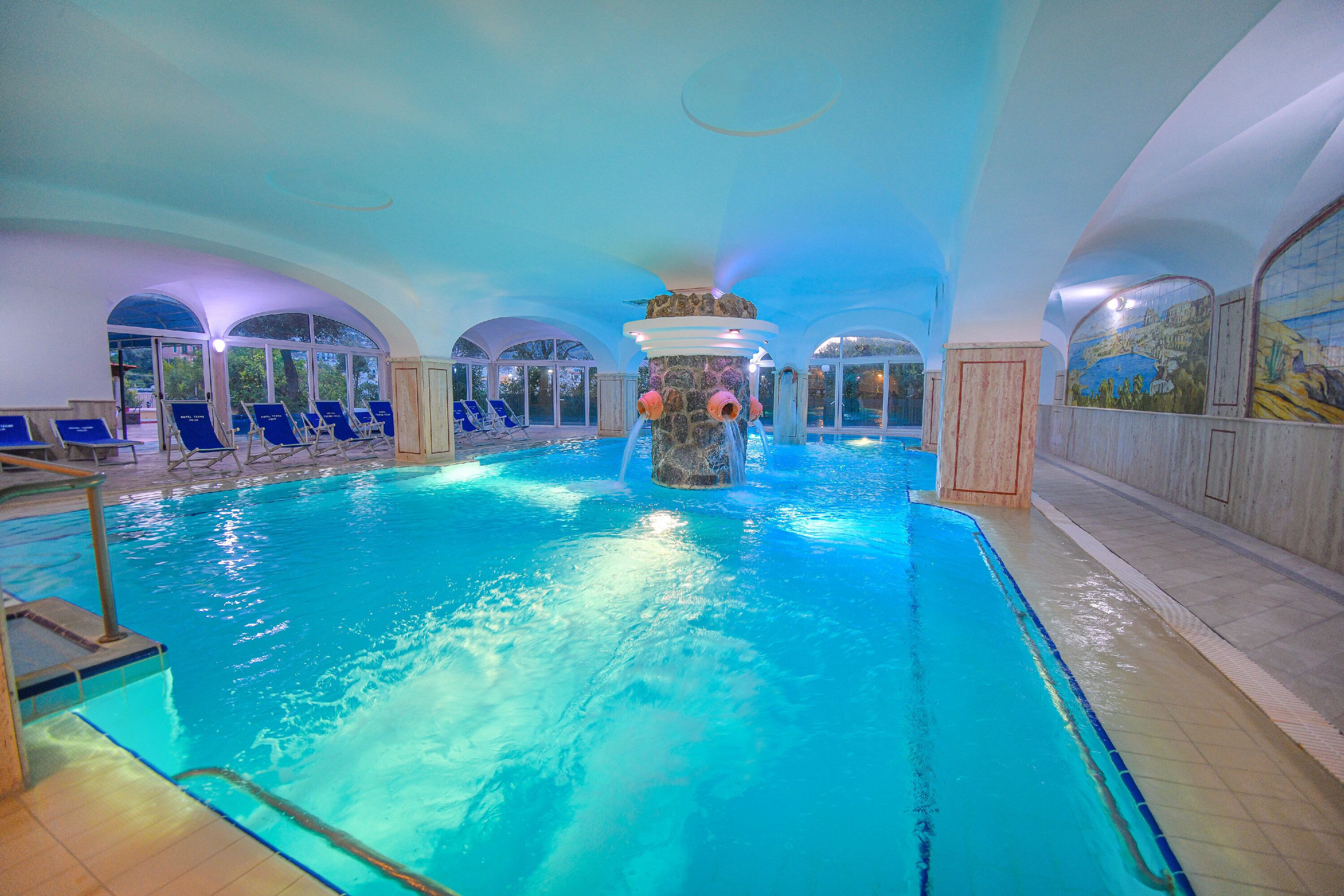 An indoor swimming pool with a central water feature, lounge chairs, and arched ceiling.