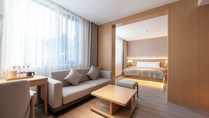 Moderni hotellihuone, jossa on sohva, pieni pöytä, sänky ja suuret ikkunat, joista avautuu näkymä kaupunkiin.