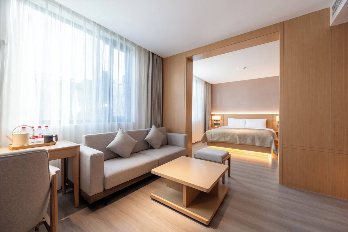 A modern hotel room with a sofa, a small table, a bed, and a view of the city through large windows.