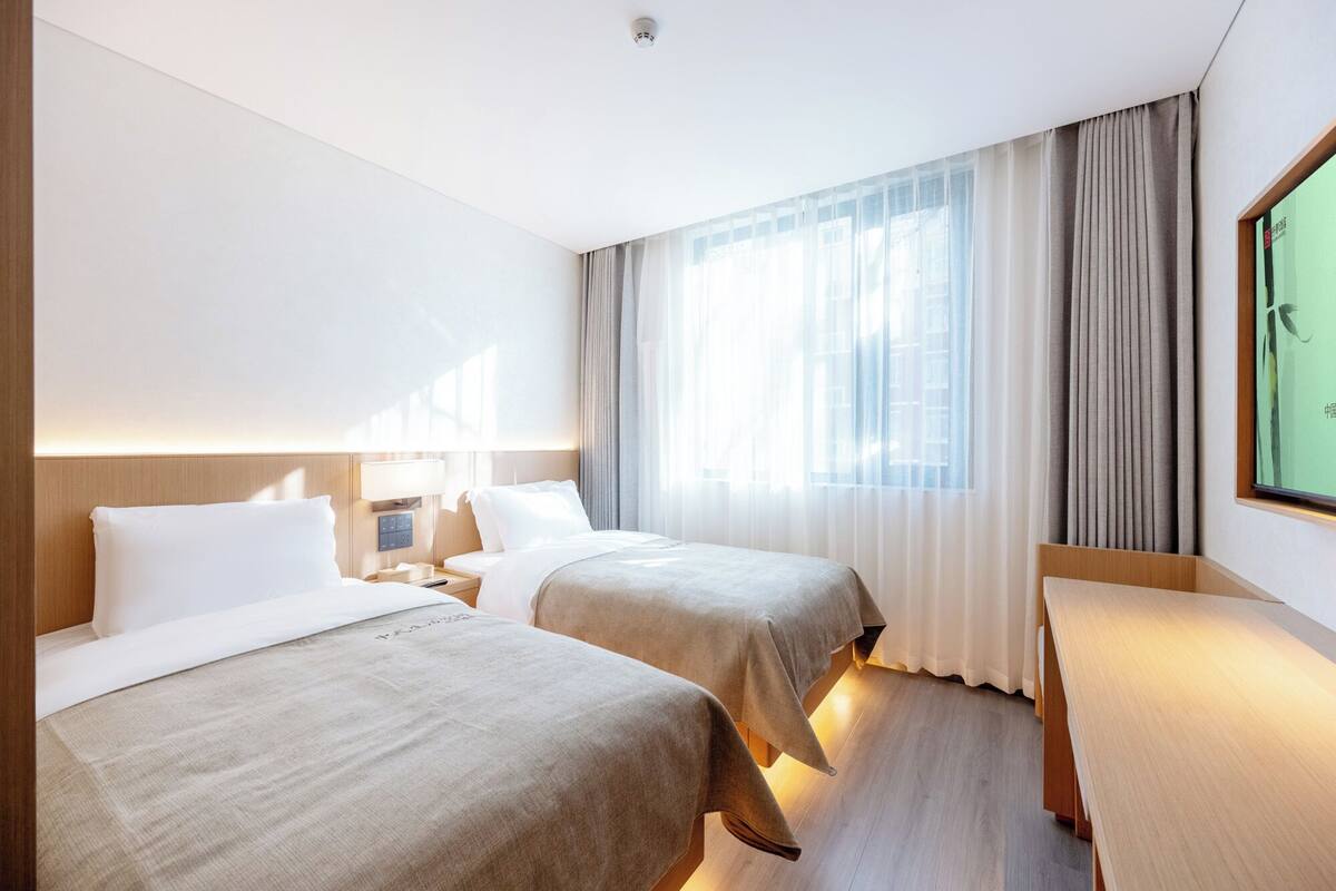A hotel room with two beds, a bedside table, a lamp, and a large window with curtains.
