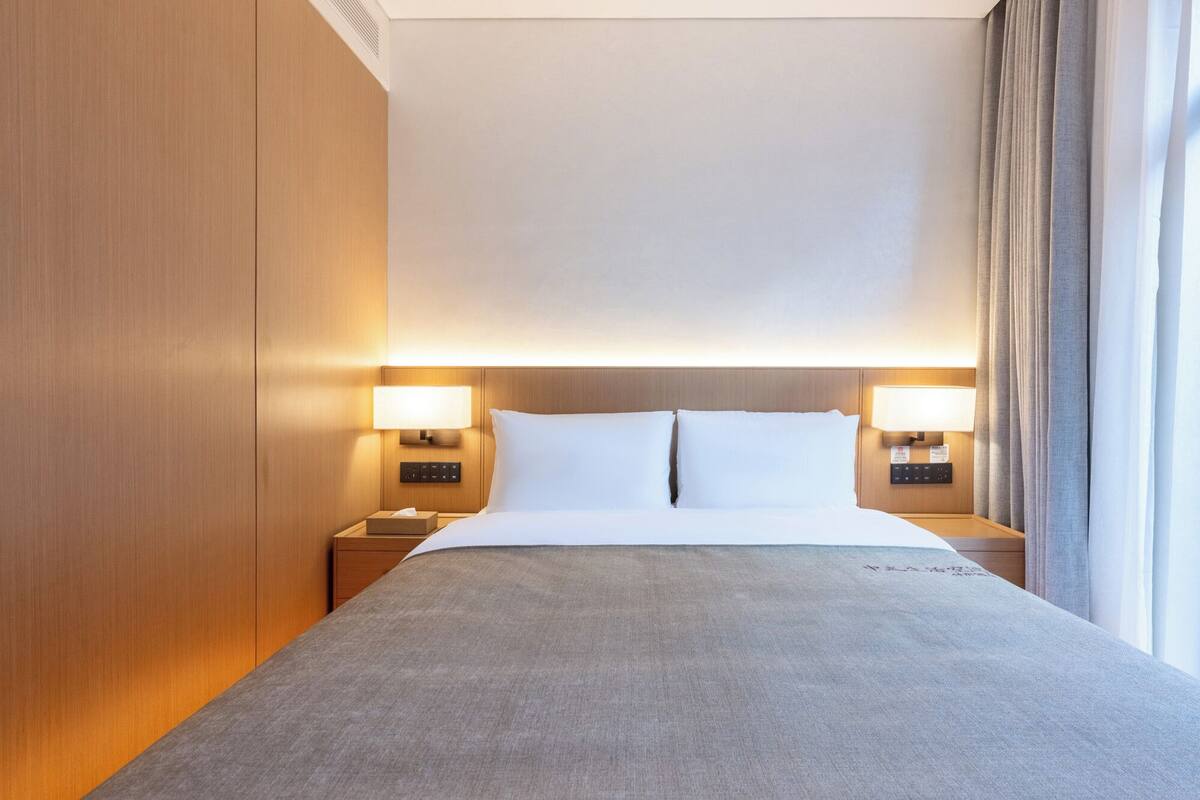 A hotel room with a bed, two bedside lamps, a nightstand, and a window with curtains.