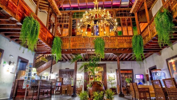 Un hall d’entrée spacieux avec un plafond haut, des poutres en bois, un lustre, des plantes suspendues et un espace salon central.