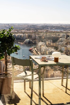 Balcón alto con una mesa, una silla, planta en maceta y vista a un paisaje urbano y una masa de agua.