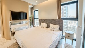 Een moderne hotelkamer met een groot bed, een flatscreen televisie aan de muur, een potplant en uitzicht op de stad door grote ramen.
