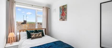 A bedroom with a bed, a nightstand, a lamp, a window with curtains, and a painting on the wall.
