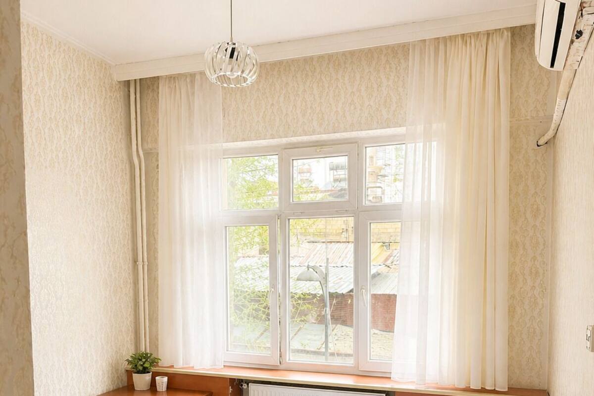 Una habitación de hotel con cama, ventana con cortinas, mesita de noche con planta y radiador.