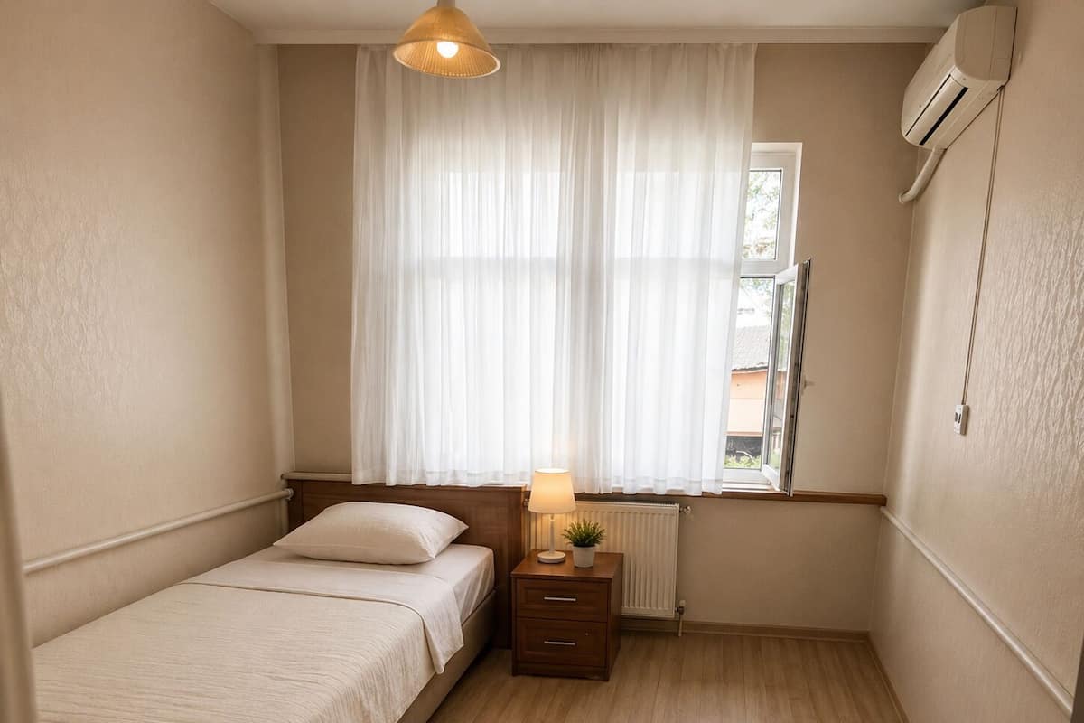 Un dormitorio pequeño y bien iluminado con una cama individual, una mesita de noche con lámpara, una ventana con cortinas traslúcidas y un equipo de aire acondicionado instalado en la pared.