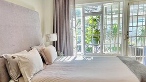 A hotel room with a large bed, a nightstand, a lamp, and a view of greenery through the open French doors. - Hout Bay Gem 61 Milner  (Cape Town)