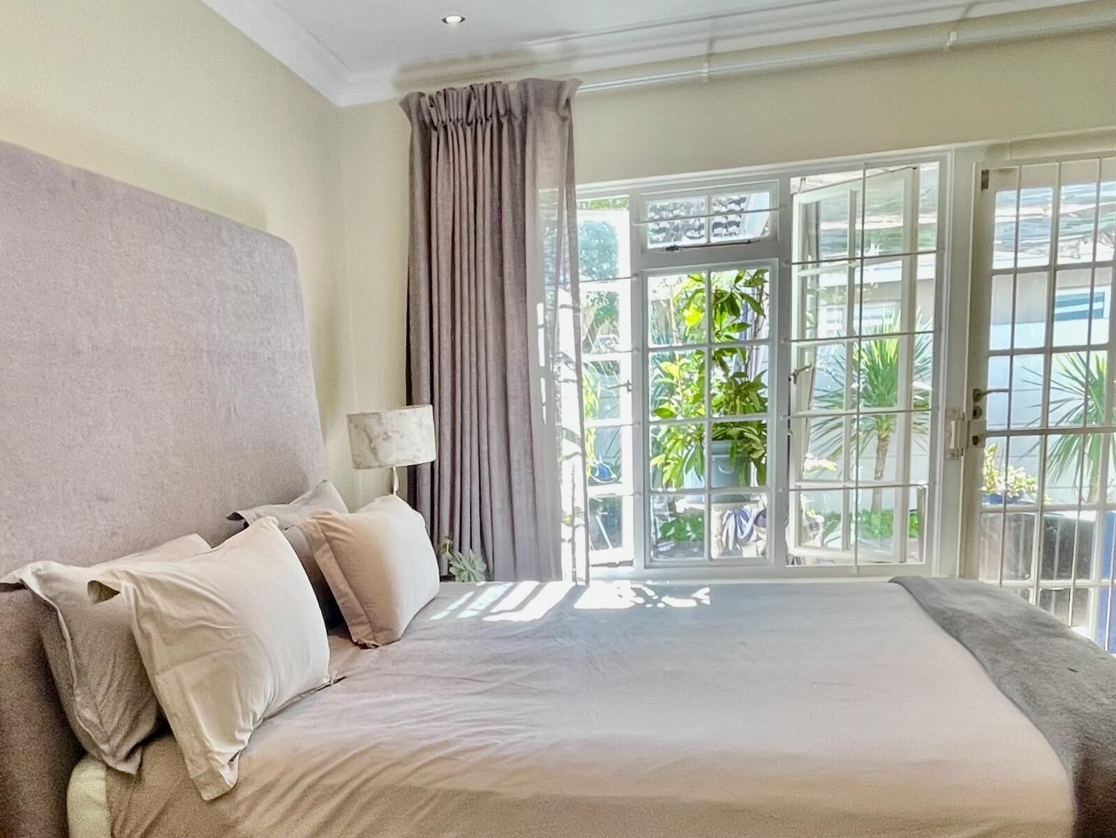 A bedroom with a large bed, a nightstand, a lamp, and a view of greenery through a glass door.