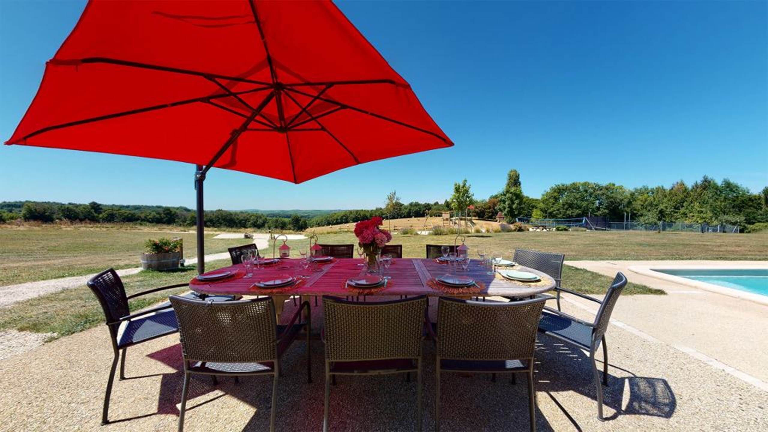 Un área de comedor al aire libre con un paraguas rojo, sillas de mimbre y una mesa preparada para cuatro.