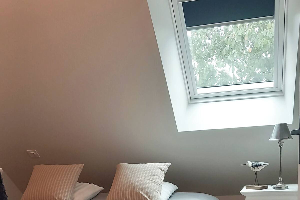 A bedroom with a sloped ceiling, a bed with striped bedding, a nightstand with a lamp, and a window with a view of trees.