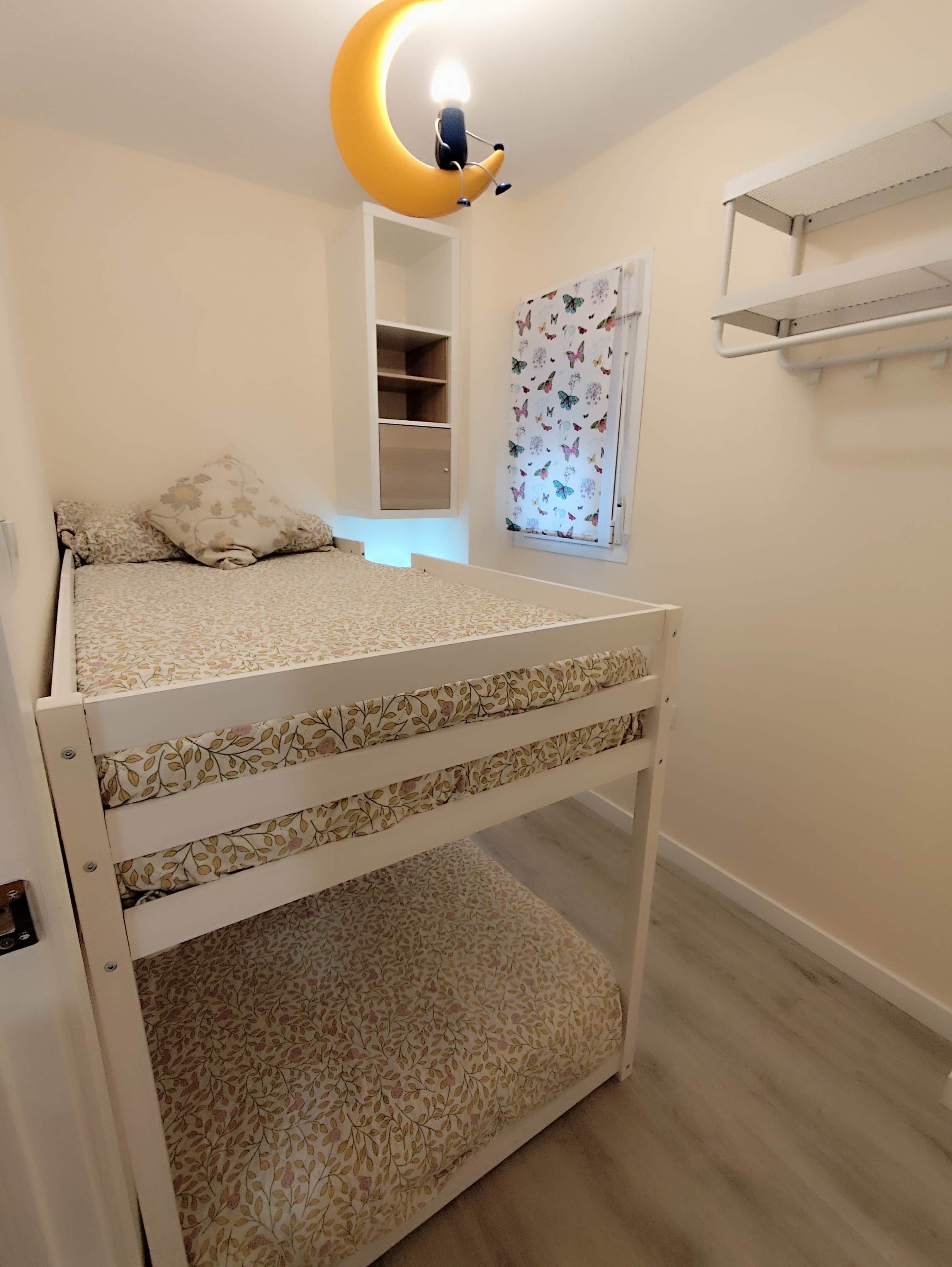 Una litera con un colchón estampado con motivos florales, una estantería fijada a la pared y una cortina con temática de mariposas.