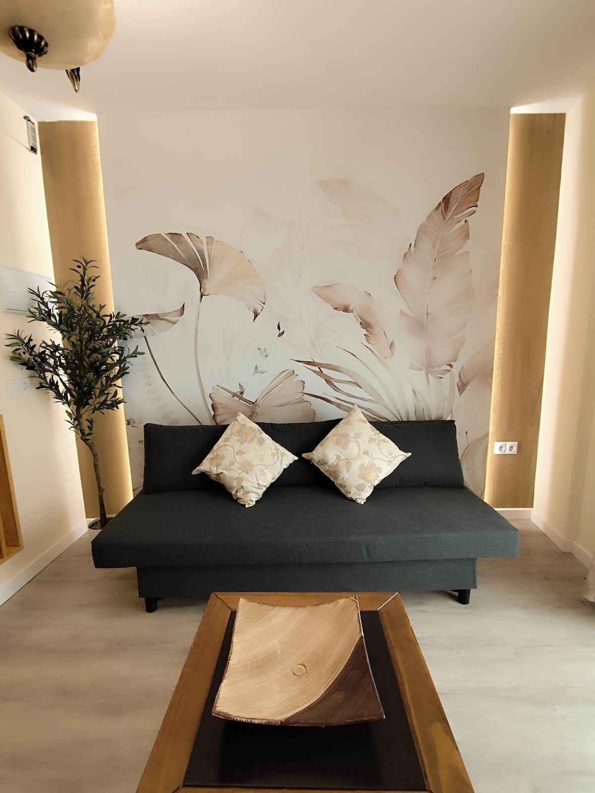 A modern living room with a dark sofa, two patterned pillows, a large abstract wall art, a wooden coffee table, and a potted plant.