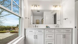 A modern bathroom with a large window, a white vanity with multiple drawers, a mirror, and a light fixture above the mirror. - Warren House 2BR, Stunning Water Views Downtown (Plymouth)