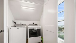 Un lavadero pequeño con dos electrodomésticos Maytag, una ventana que da a la calle y edificios, y una puerta que conduce a otra habitación.