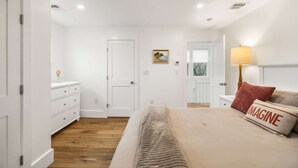 Un dormitorio con cama, mesita de noche, lámpara, cómoda, puerta y un cuadro en la pared.
