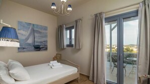 A bedroom with a bed, a painting, a chandelier, a window, and a balcony with a table and chairs. - Harites Home – Sea View Villa A | Peaceful Island Escape with Stunning Views (Milos)
