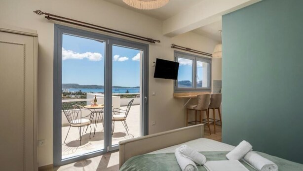 A hotel room with a bed, a TV, a balcony with chairs, and a view of the sea. - Harites Home – Sea View Villa A | Peaceful Island Escape with Stunning Views (Milos)