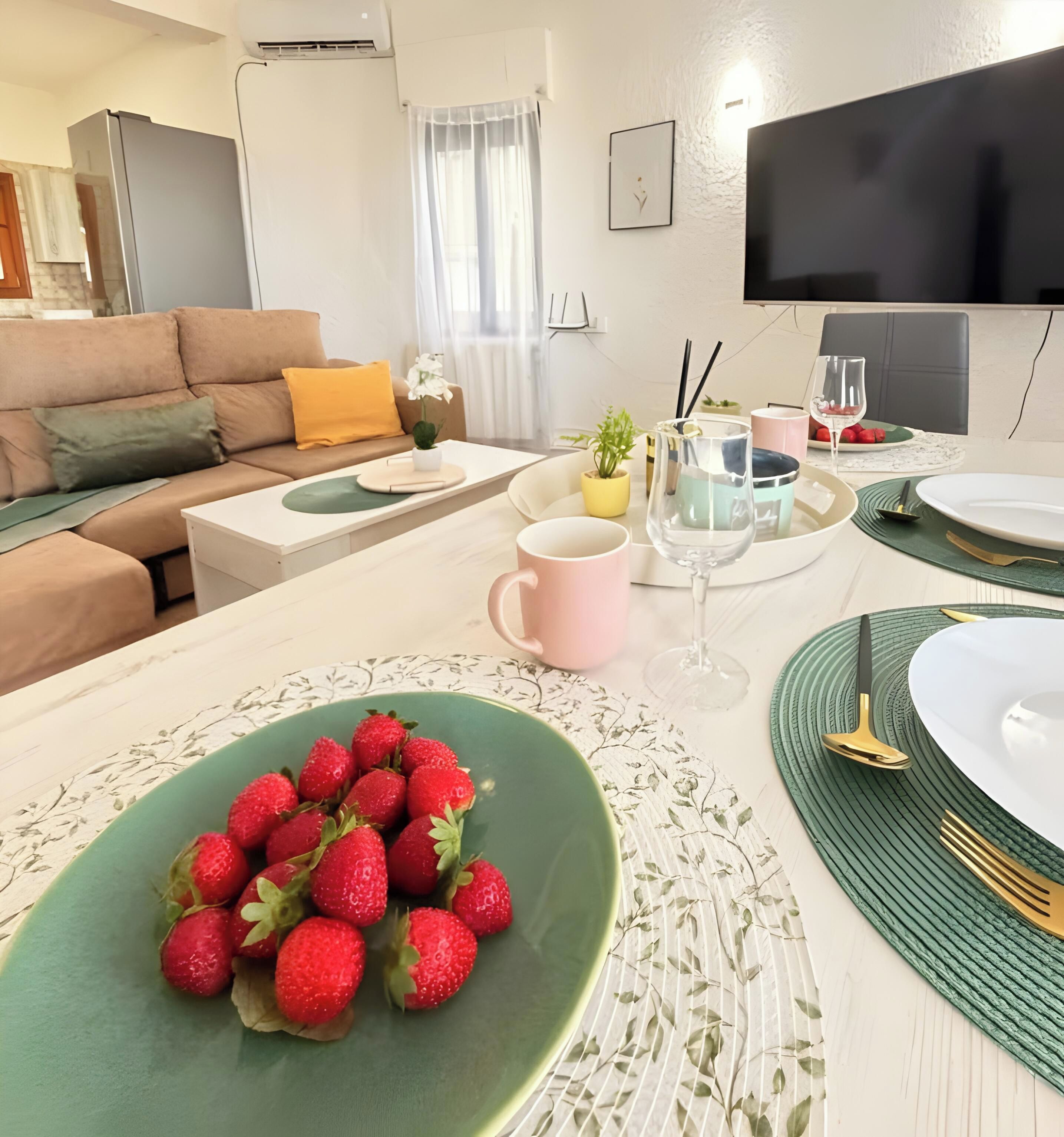 A modern living room with a sofa, a dining table set with plates, cutlery, and a bowl of strawberries.